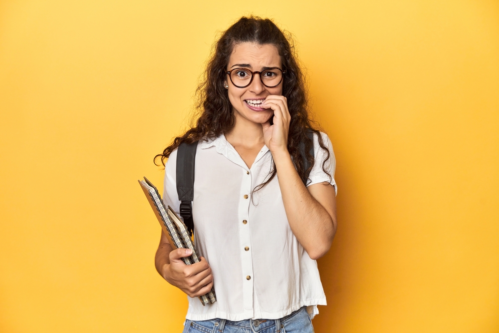 Studentessa con i libri in mano si morde le dita dalla preoccupazione. su sfondo giallo